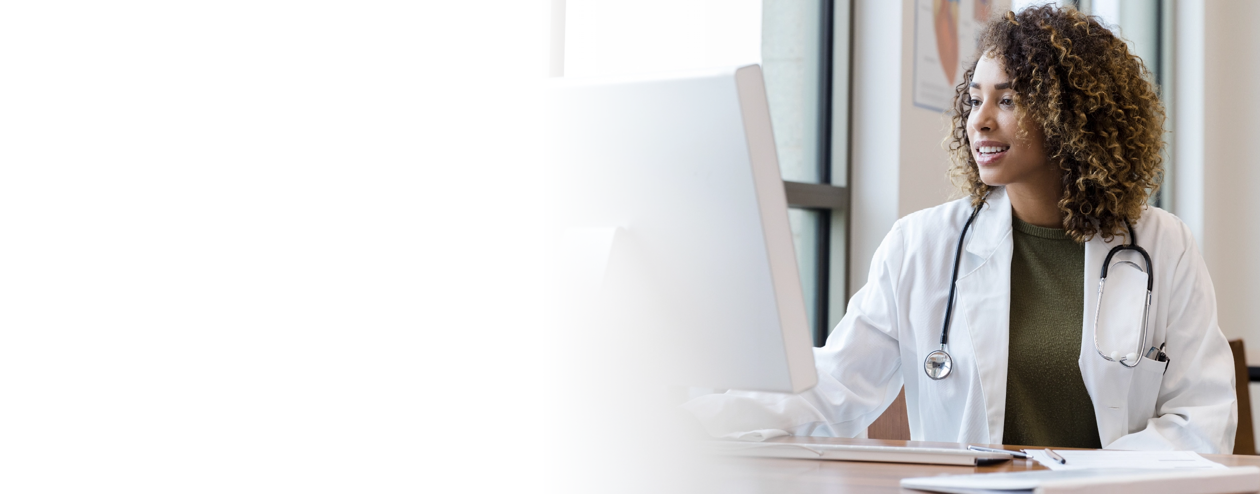 Female healthcare professional wearing white lab coat with stethoscope looking at computer screen.