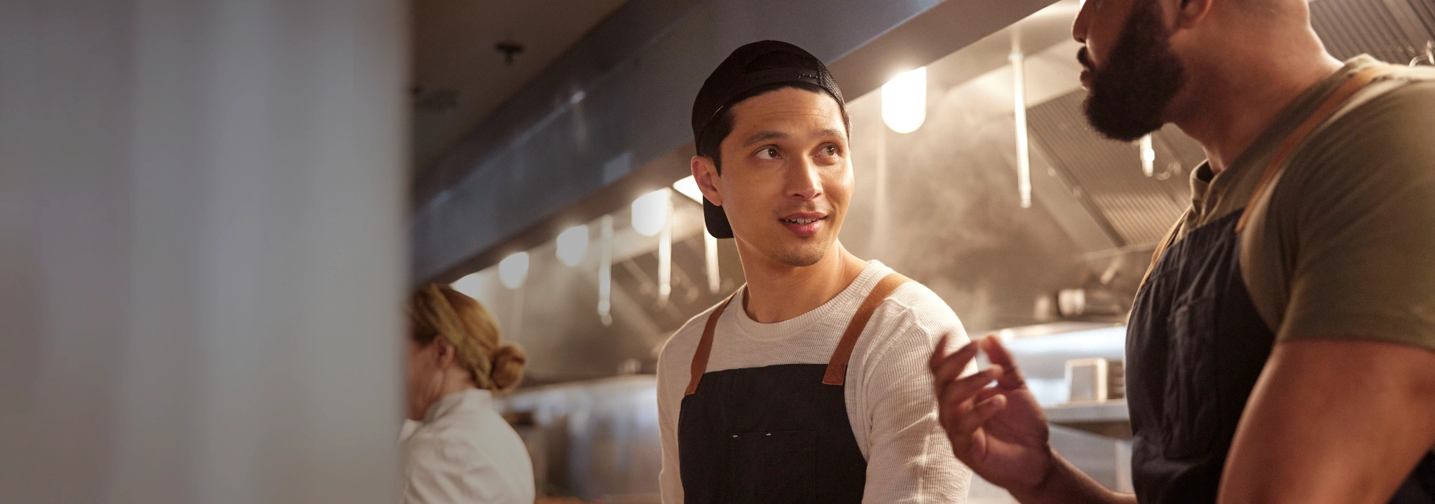 Two men in aprons talking in a kitchen